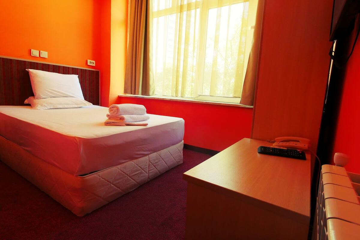 A hotel room with a bed, a bedside table with a remote control, a phone, and a window with curtains.
