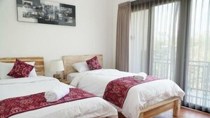 Ein Hotelzimmer mit zwei Betten, jeweils mit weißer Bettwäsche und roten Zierkissen. Dazwischen befindet sich eine Holzbank, und ein großes Fenster mit Vorhängen.