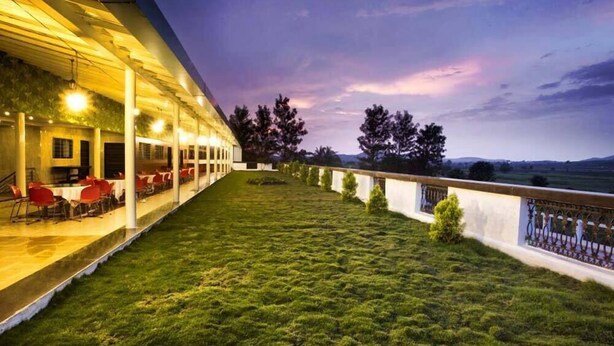 A covered outdoor seating area with tables and chairs, a grassy lawn, and a view of the landscape. - GP Farm (Nashik)