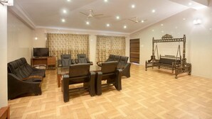 A spacious living room with a wooden floor, a glass coffee table surrounded by leather chairs, a television, and a swing bed against the wall. - GP Farm (Nashik)