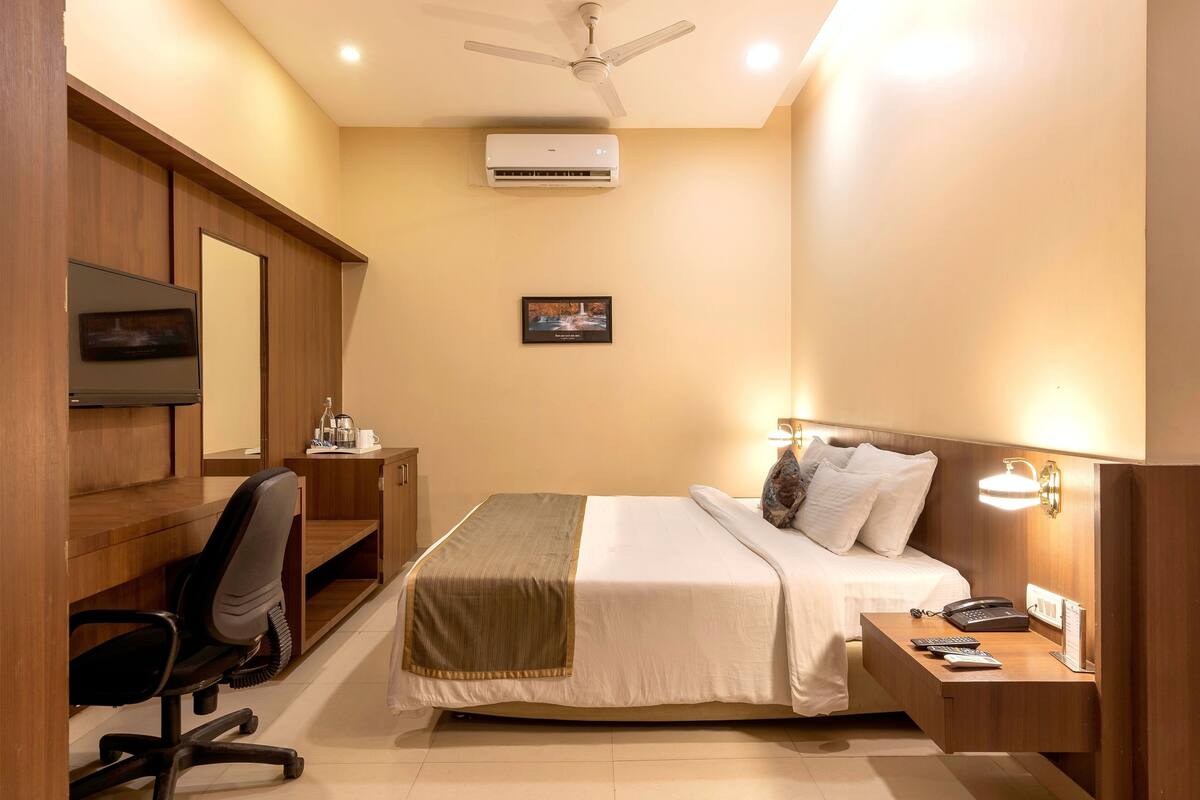 A hotel room with a bed, desk, chair, bedside table, lamp, and an air conditioning unit.