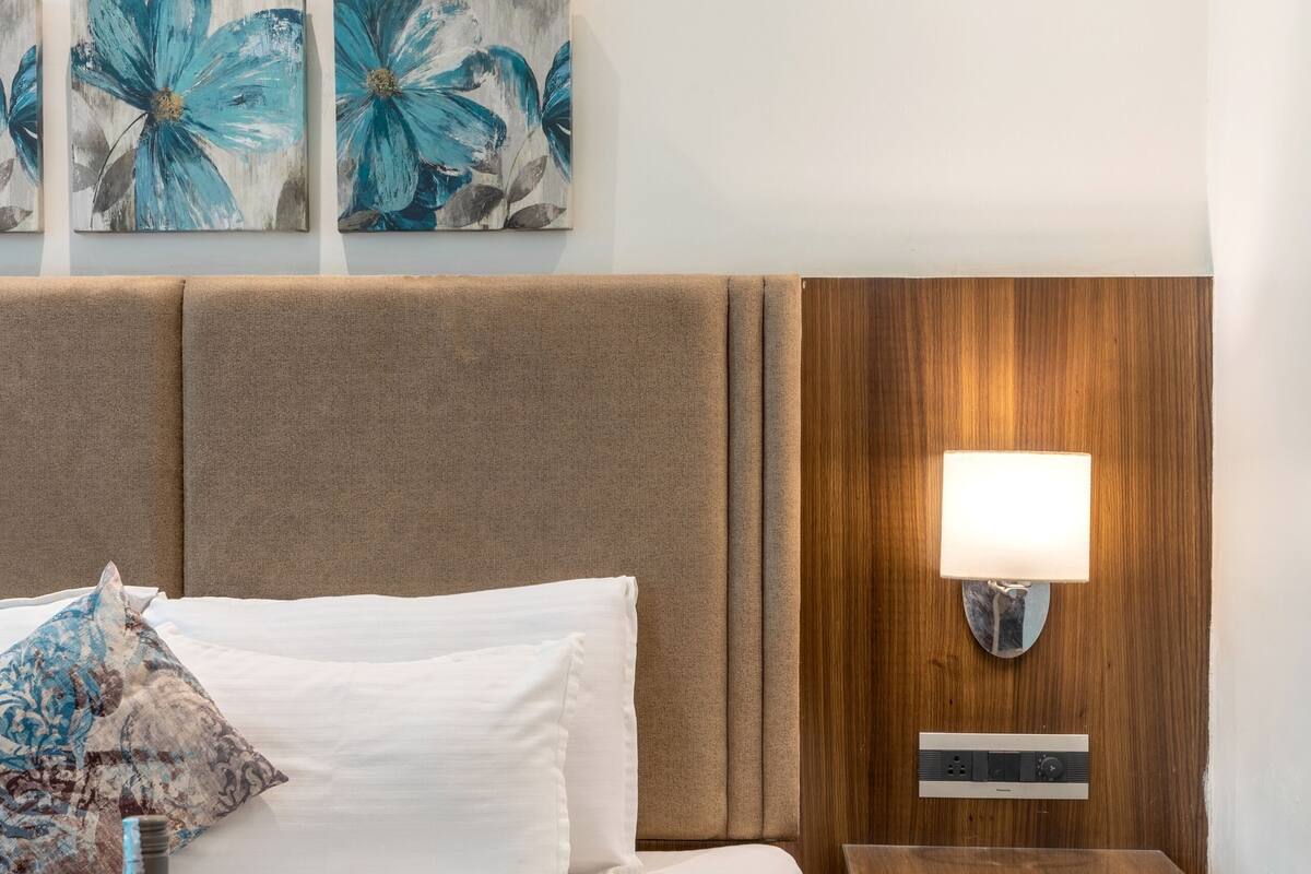 A hotel room with a bed, a bedside table with a lamp, a tray with wine glasses and a bottle, and three floral paintings on the wall.