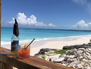 Une bouteille de bière et un cocktail, posés sur une rambarde en bois surplombant une plage aux eaux bleues et cristallines, avec une terre lointaine à l’horizon.