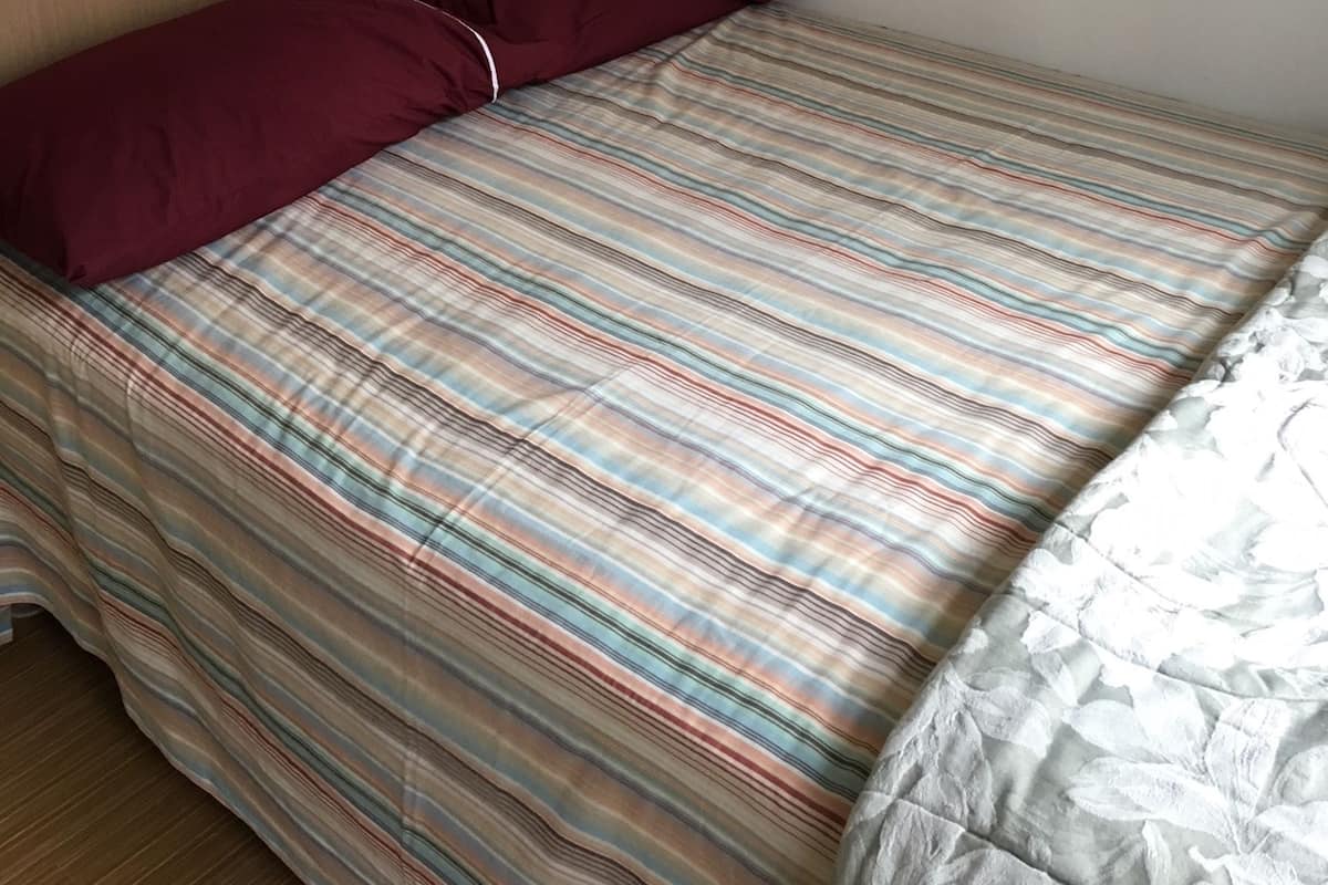 A single bed with a striped bedspread and two pillows, one red and one white, in a room with wooden paneling and a plain wall.
