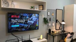 A modern living room with a mounted TV displaying Netflix, a coffee table with a lamp, a chair, and a shelf with decorative items.