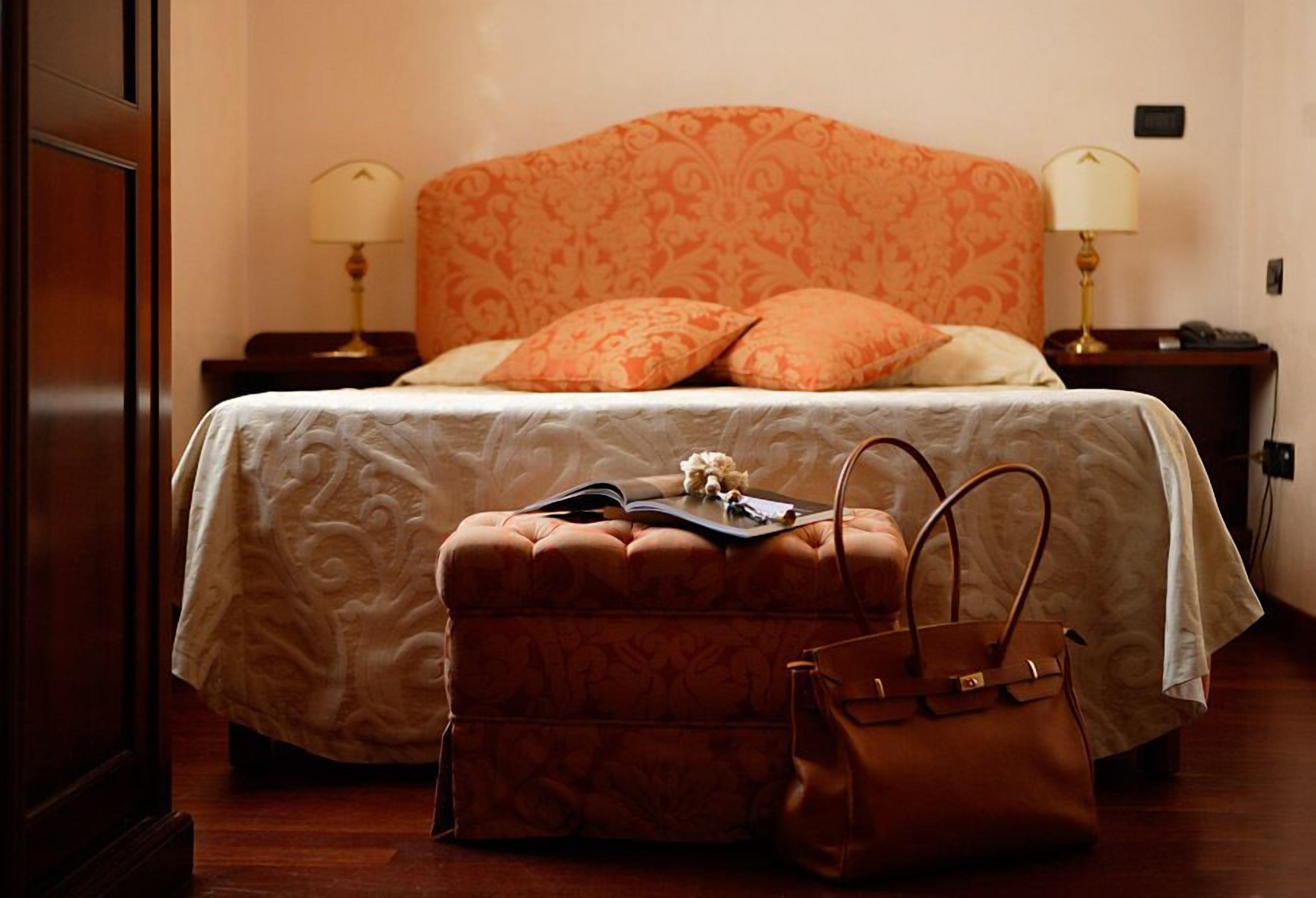 Ein ordentlich bezogenes Bett mit einem gemusterten Kopfteil, zwei Nachttischlampen, ein kleiner Tisch mit einem Buch und einer Handtasche, sowie ein Holzboden.