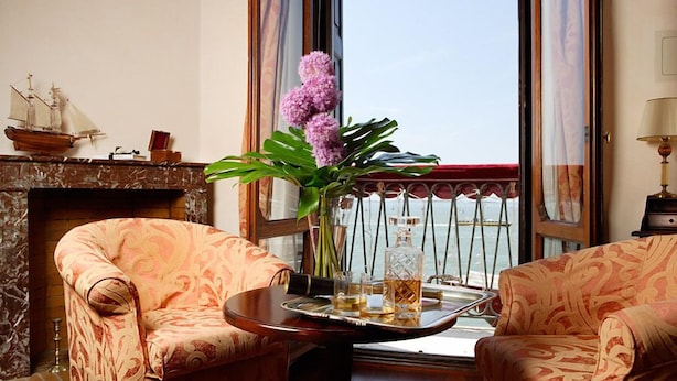 Une pièce avec deux fauteuils à motifs, une petite table sur laquelle est posé un vase de fleurs, une maquette de bateau, et une vue sur la mer depuis un balcon.