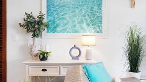 Una habitación con pared blanca, un cuadro enmarco con imagen de agua, una cruz de madera, un escritorio blanco con lámpara, un florero con planta y una planta en maceta.