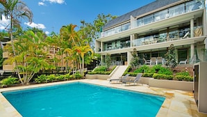Un edificio moderno de dos pisos con piscina, mobiliario exterior y abundante vegetación.