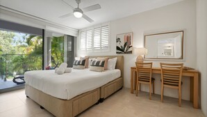 Un dormitorio con cama, escritorio, dos sillas, ventilador de techo y balcón con vista a árboles.