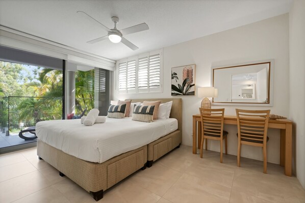 A bedroom with a bed, a desk, two chairs, a ceiling fan, and a balcony with a view of trees.