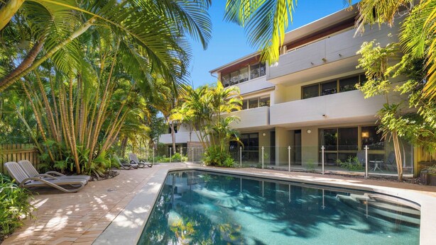Uma área de piscina moderna com espreguiçadeiras, palmeiras e um edifício de dois andares com grandes janelas.