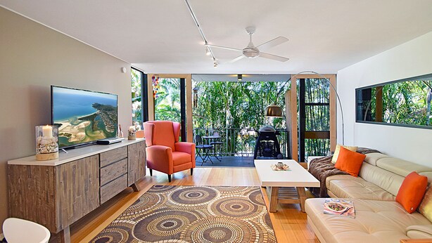 A modern living room with a TV, a red armchair, a beige sofa, and a patterned rug. - Ludlow Apartments (Noosa Heads)