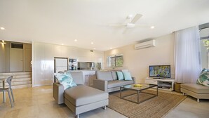 A modern living room with a sofa, coffee table, and TV. There is a kitchen in the background with cabinets and a refrigerator. - Outlook (Noosa Heads)