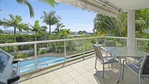 A balcony with a glass table and chairs overlooking a swimming pool, surrounded by palm trees and a view of the ocean. - Outlook (Noosa Heads)