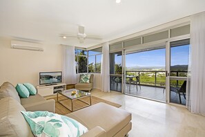 A modern living room with a sofa, coffee table, and TV, leading to a balcony with a view of the landscape. - Outlook (Noosa Heads)