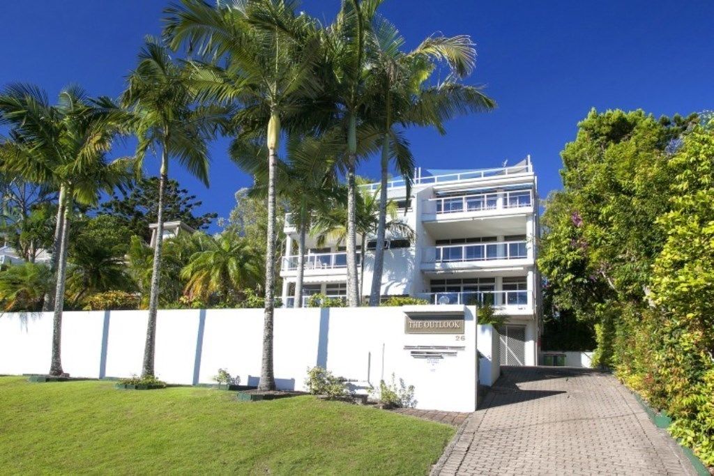 Un bâtiment moderne à la façade blanche, agrémenté de palmiers et doté d’un chemin pavé menant à l’entrée.