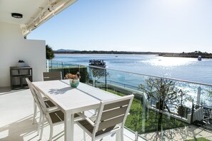 Un balcón con mesa y sillas blancas, con vista a un cuerpo de agua y barcos.