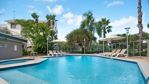 A swimming pool with lounge chairs and umbrellas, surrounded by palm trees and a building with a grey facade.