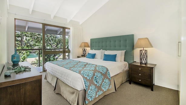 A bedroom with a large bed, two bedside tables, a dresser, and a window with a view of greenery. - Kananda (Noosa Heads)