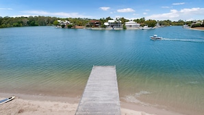 A wooden dock leading to a sandy beach with calm water, a boat in the distance, and houses on the opposite shore. - Weyba Quays (Noosa Heads)