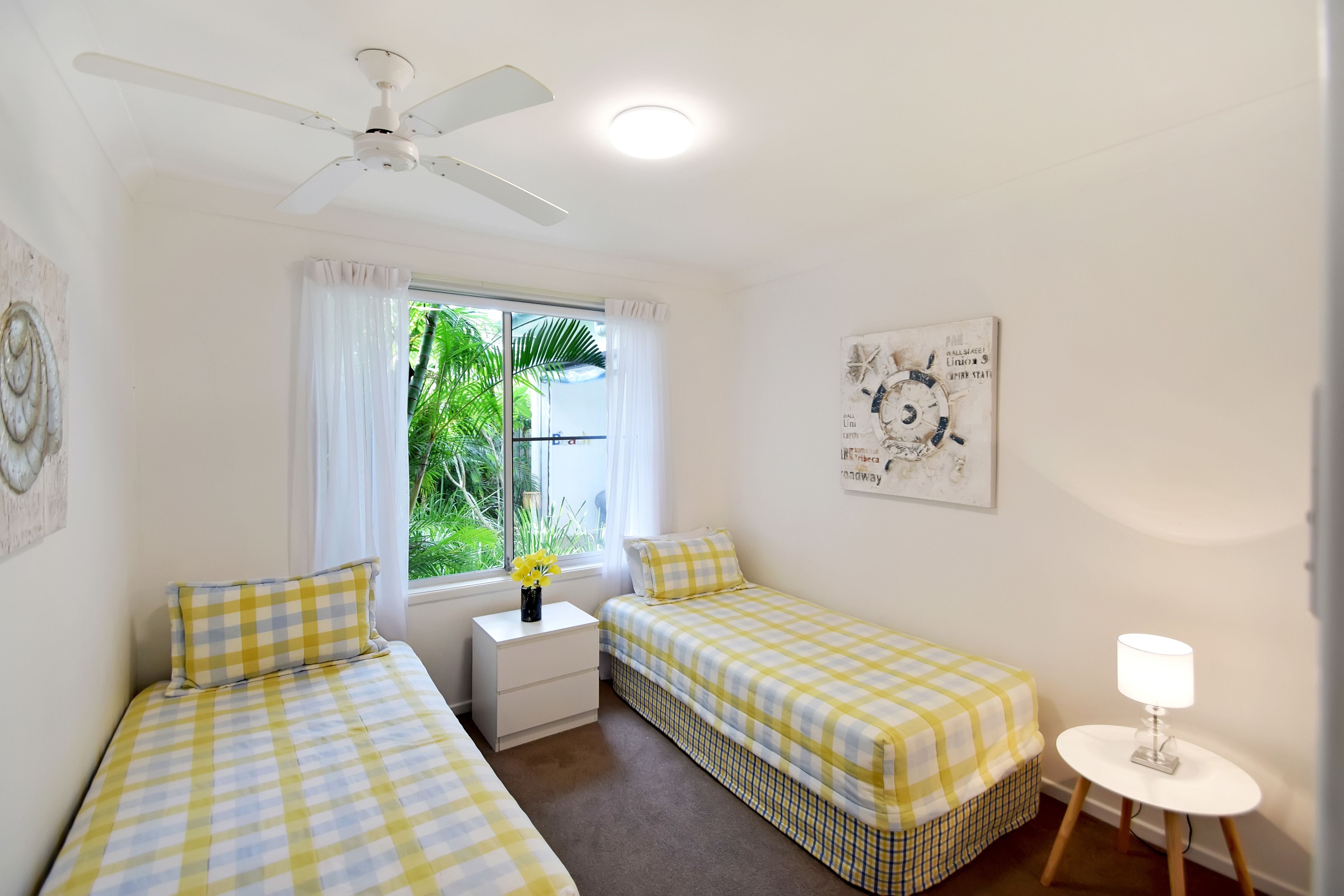 A bedroom with two single beds, a ceiling fan, a nightstand, a small table with a lamp, and a window with curtains.