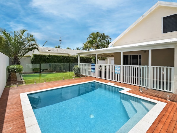 A rectangular swimming pool with a wooden deck surrounding it, a covered patio area with a white railing, and a well-maintained lawn in the background.