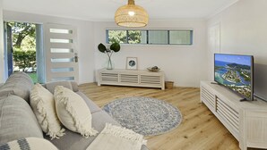 Ein modernes Wohnzimmer mit einem grauen Sofa, einem runden, blau-weißen Teppich, einem weißen Konsolentisch und einem Flachbildfernseher sowie einem großen Fenster mit Jalousien.