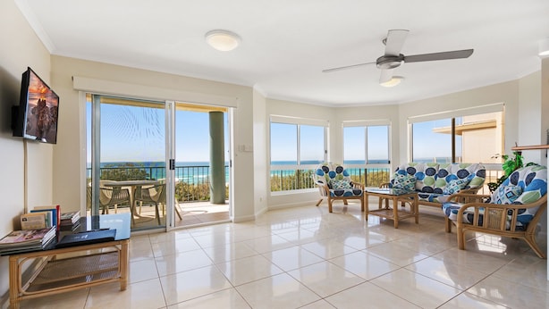 A spacious living area with a balcony overlooking the ocean, furnished with wicker chairs and a table, and a ceiling fan. - Pacific Waves (Sunrise Beach)