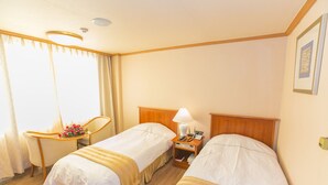 Habitación de hotel con dos camas, un escritorio con silla, una lámpara y una pequeña mesa con un arreglo floral.