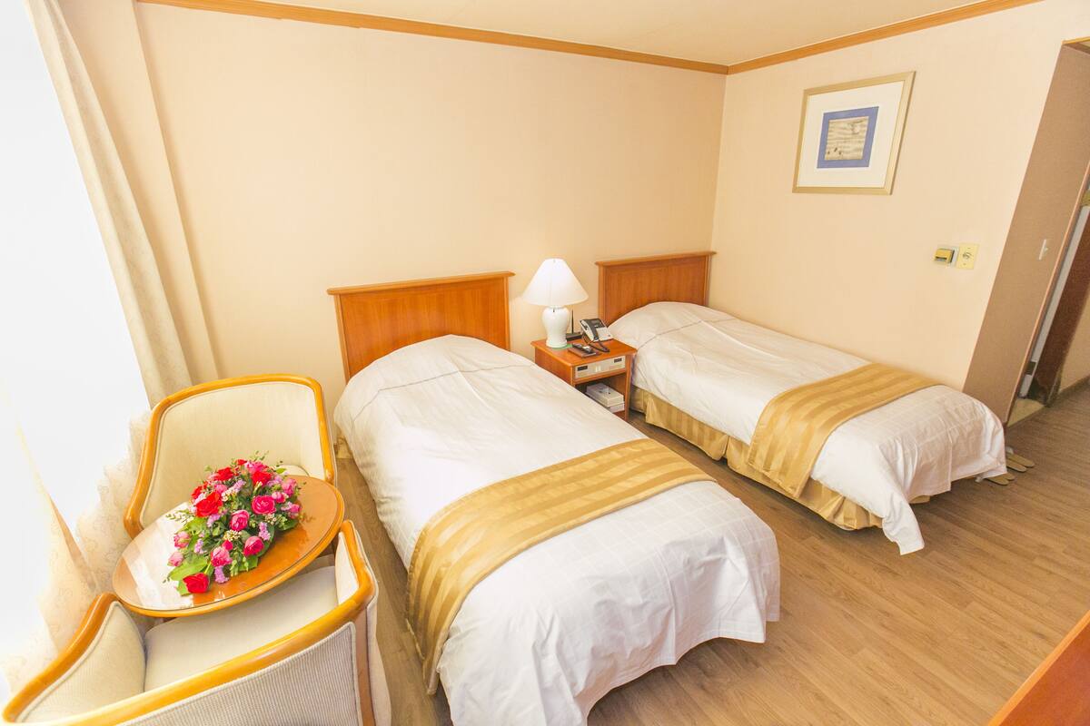 A hotel room with two beds, a chair with a floral arrangement, a bedside table with a lamp, and a framed picture on the wall.