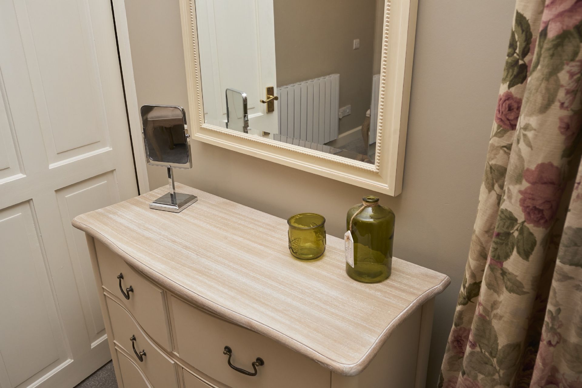 A bathroom with a wooden vanity, a mirror, a green glass bottle, and a floral curtain.
