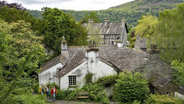A traditional stone house with a thatched roof, surrounded by lush greenery and hills in the background. - Old Bakers Cottage, ground floor centrally located cottage in the heart of Grasmere. (Ambleside)