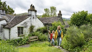 A white stone house with a thatched roof, surrounded by lush greenery and a stone pathway. - Old Bakers Cottage, ground floor centrally located cottage in the heart of Grasmere. (Ambleside)