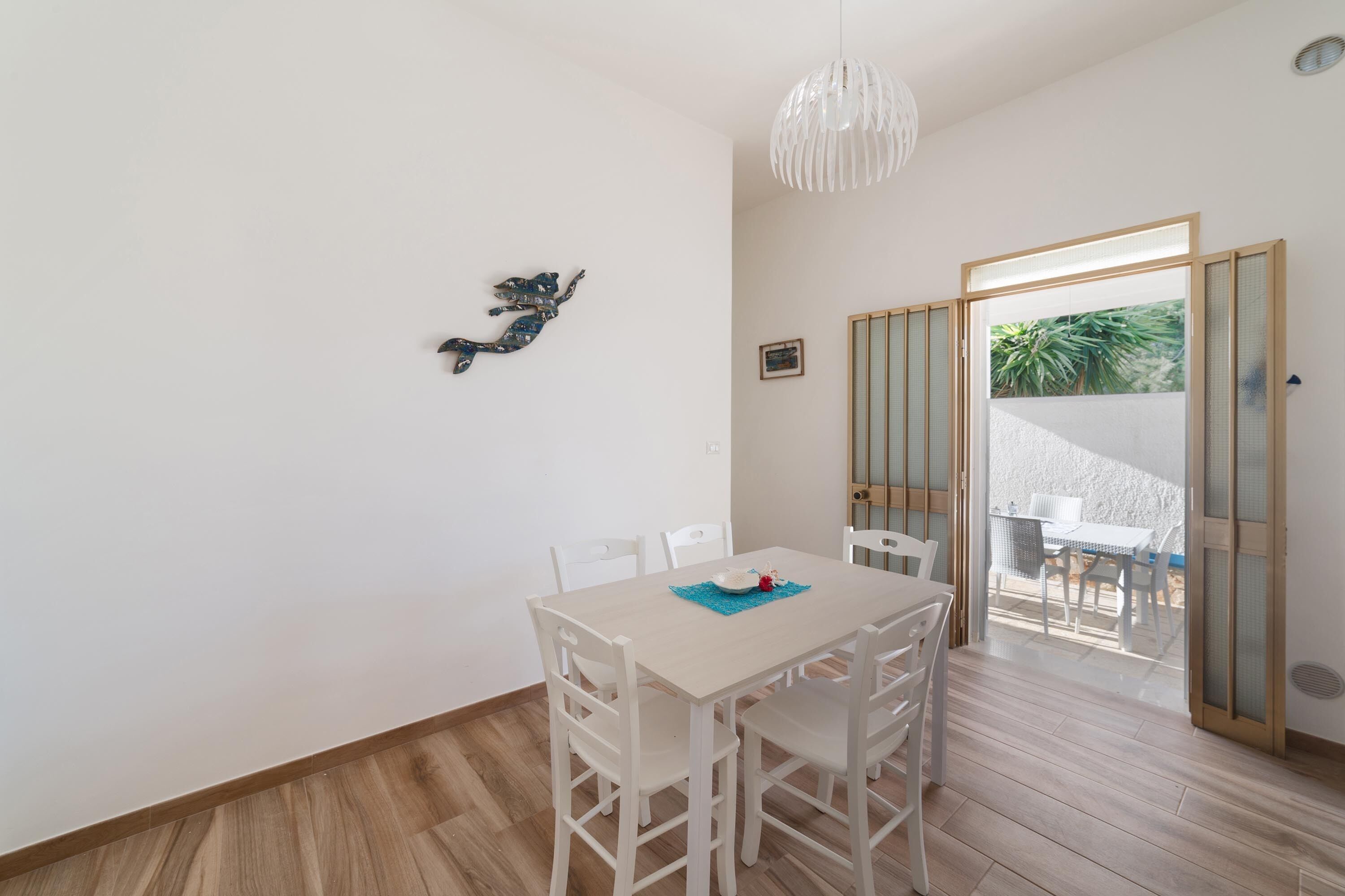 Un comedor con una mesa y sillas blancas, un adorno en la pared, y vistas a un patio exterior a través de un ventanal.