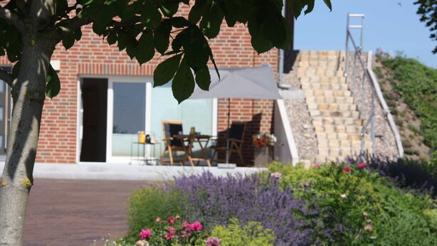 Ein modernes Gebäude mit Backsteinfassade, einer überdachten Außenbereich-Sitzgelegenheit und einem angelegten Garten mit verschiedenen Pflanzen und Blumen.