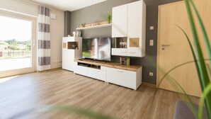 Ein modernes Wohnzimmer mit Flachbildfernseher, Holzboden und einer Schiebeglastür, die auf einen Balkon führt.