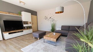 Ein modernes Wohnzimmer mit Flachbildfernseher, Holztisch, grauem Sofa und Wanduhr.
