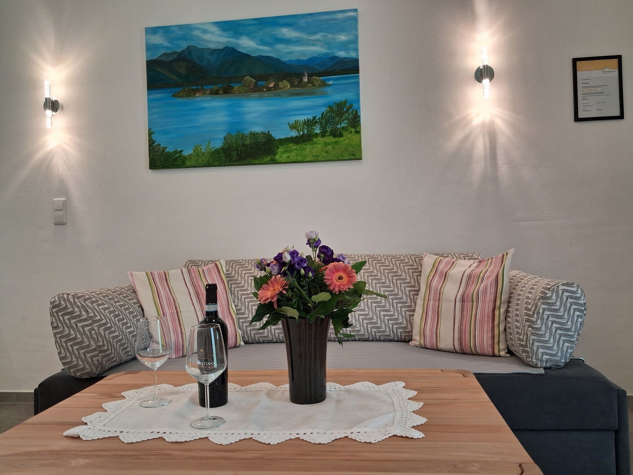 A living room with a sofa, a table with a vase of flowers, a wine bottle, and two wall-mounted lights.