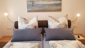 Une chambre à coucher avec un lit, deux oreillers, une table de chevet, une télécommande et un tableau représentant une scène de plage.