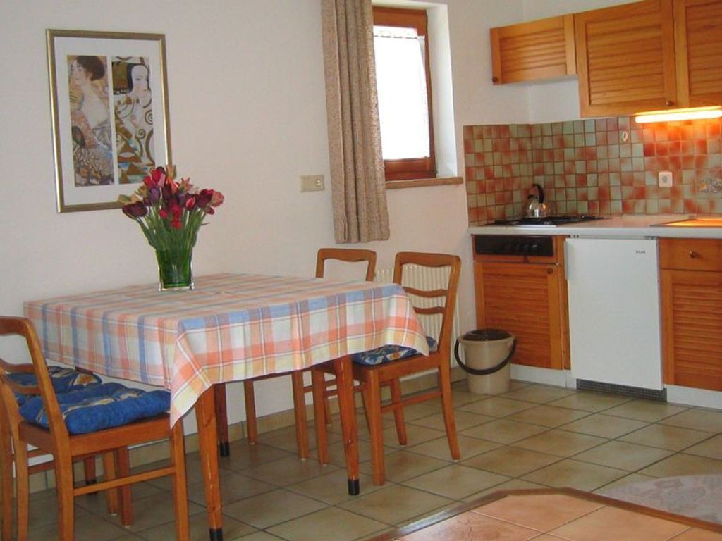 Ein Essbereich mit Tisch, Stühlen, einem Blumenstrauß, einer Küche mit Schränken, einem Kühlschrank und einer gefliesten Spritzwand.
