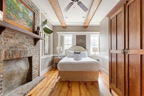 A bedroom with a brick fireplace, a large bed, wooden ceiling beams, and a view of the outside through a window. - The Marquee On King & Historic Market! (Charleston)