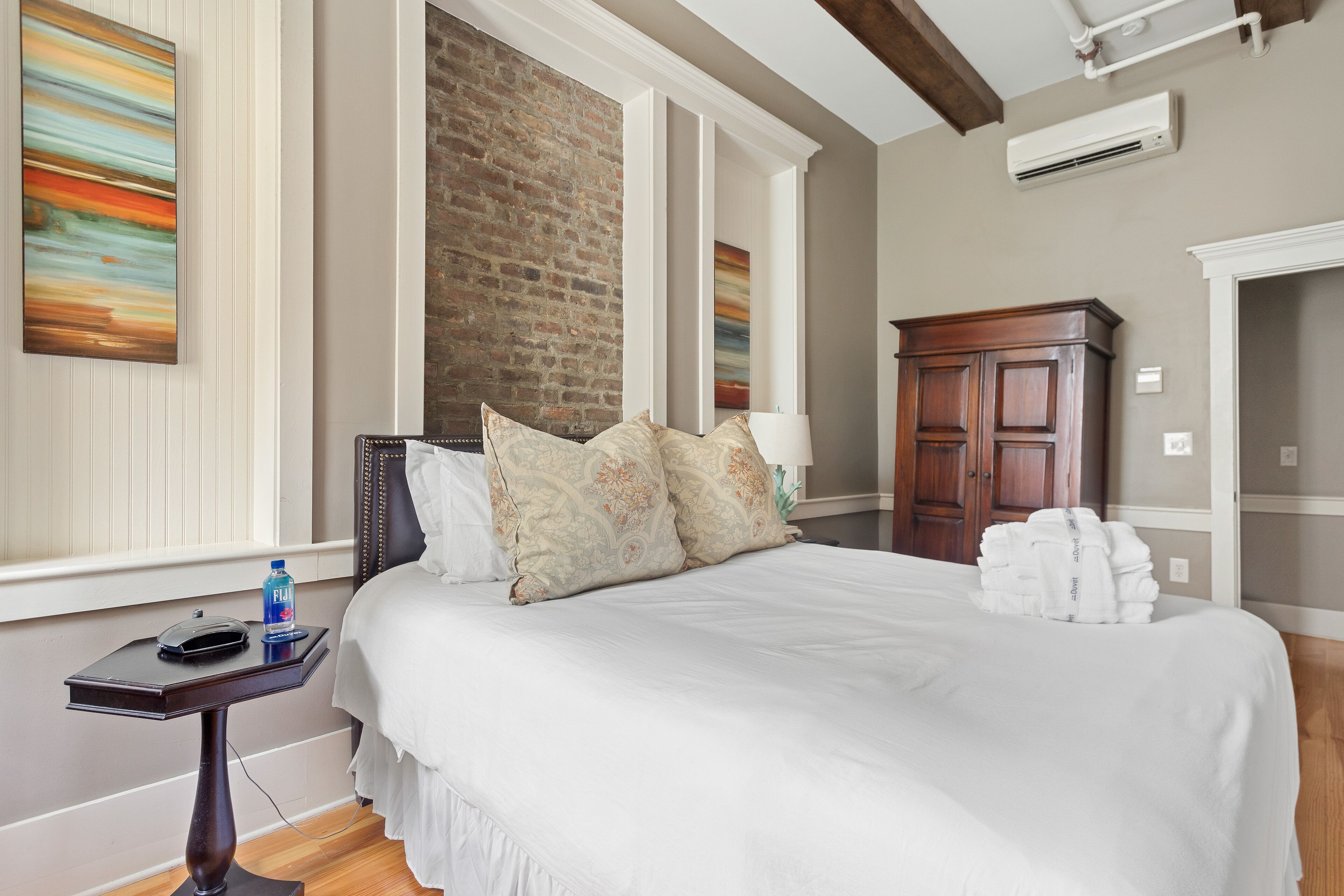 A neatly made bed with white linens, a bedside table with a lamp, and a brick wall behind the bed.