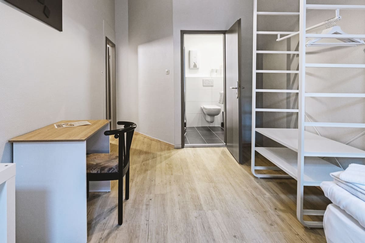 Quarto de hotel com uma secretária de madeira, uma cadeira, uma cama, um guarda-roupa e um banheiro visível através de uma porta aberta.