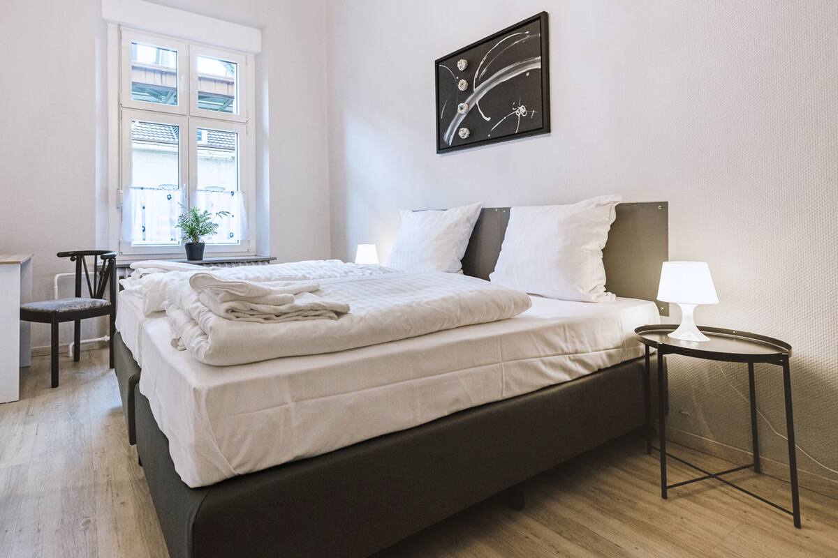 Cama bem arrumada com edredom branco e travesseiros, mesa de cabeceira com abajur, pequena escrivaninha com cadeira e janela com persianas.