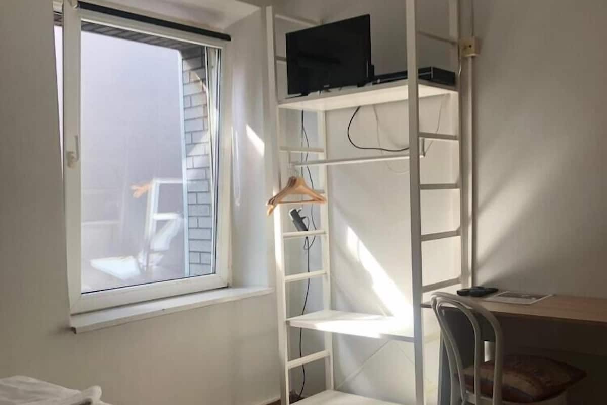 Um quarto com cama, escrivaninha com cadeira, televisão numa prateleira e janela.