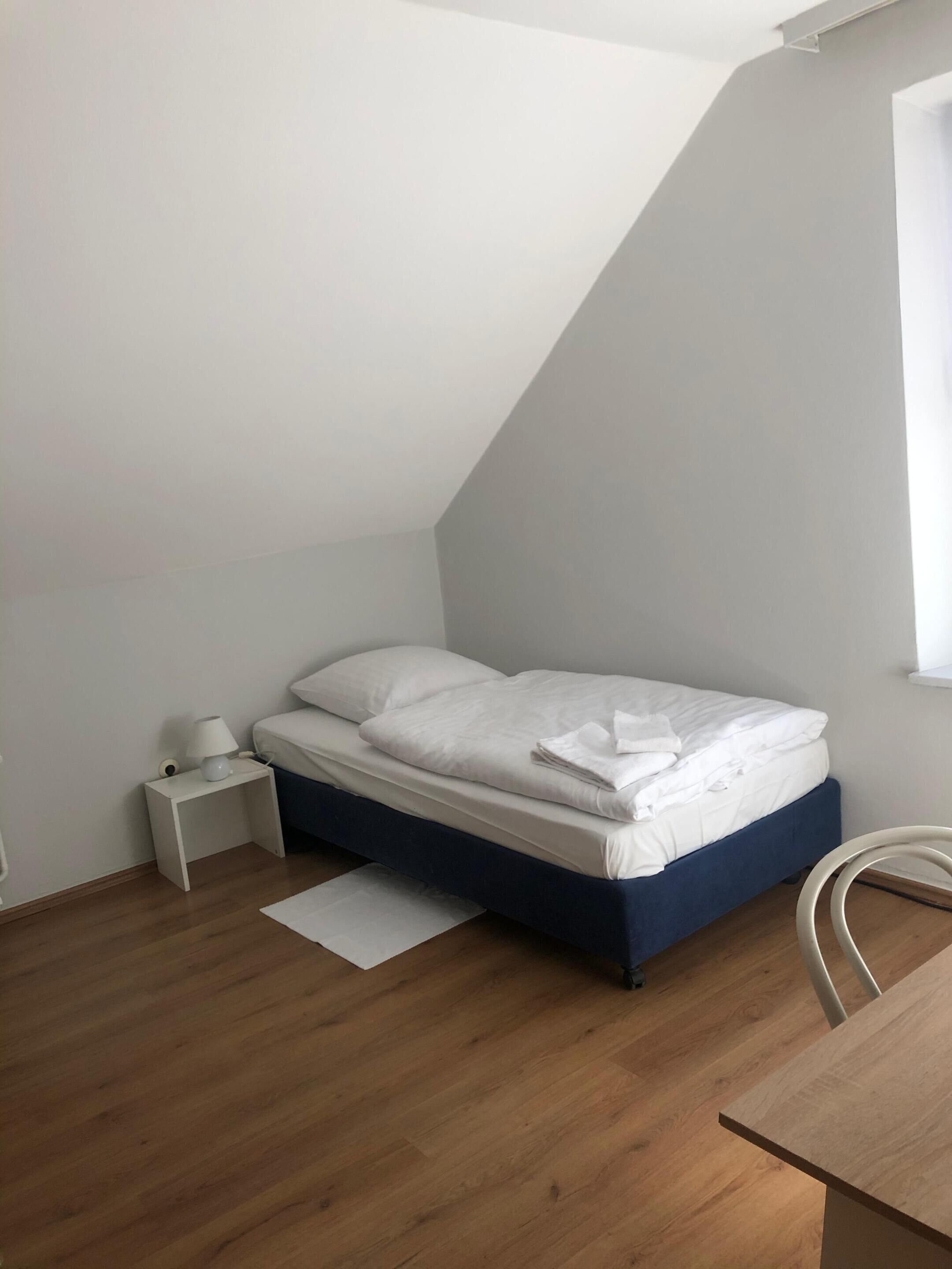 A small, neatly arranged bedroom with a single bed, a bedside table with a lamp, and a chair.