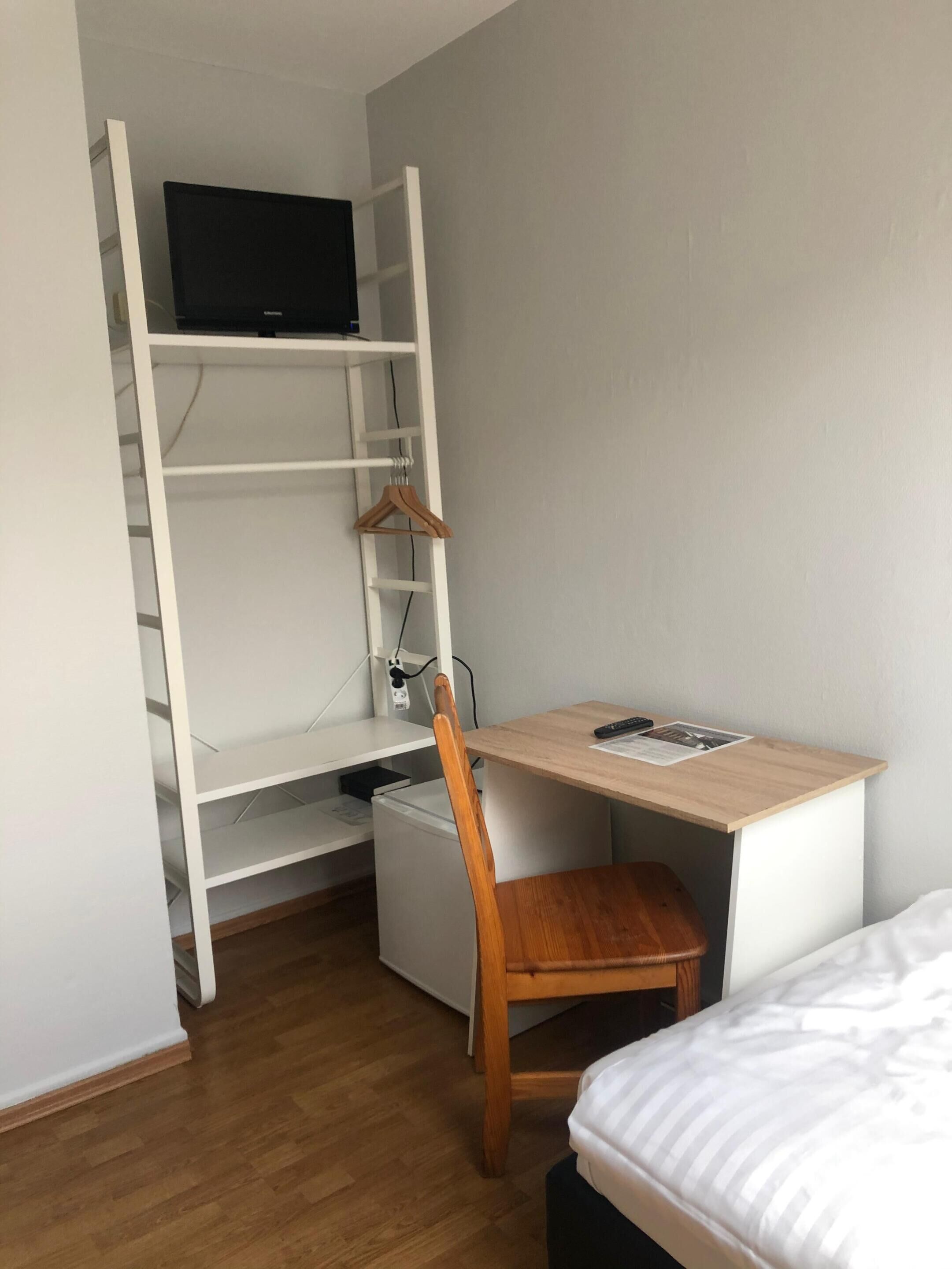 Une petite pièce comprenant un lit, un bureau avec une chaise, un téléviseur posé sur une étagère et un petit réfrigérateur.