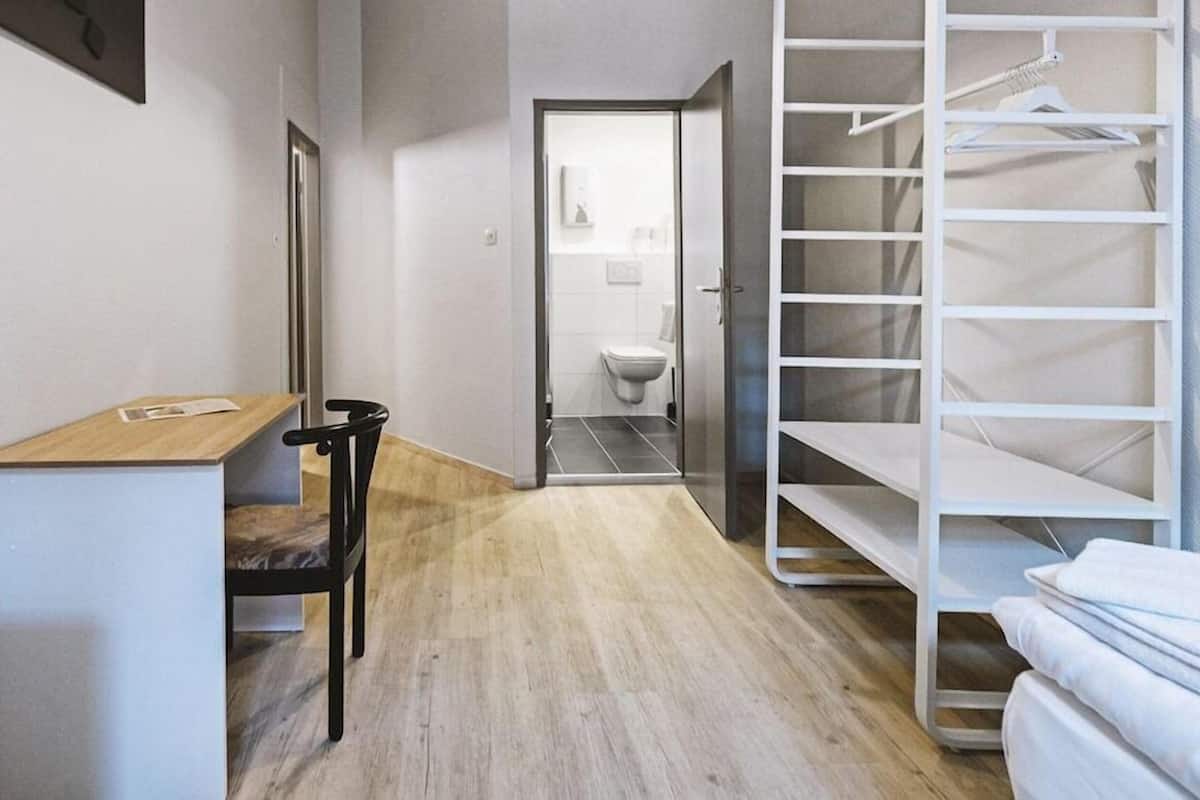 Quarto de hotel com escrivaninha de madeira, uma cadeira, uma cama, um guarda-roupa com prateleiras e uma casa de banho visível através de uma porta aberta.