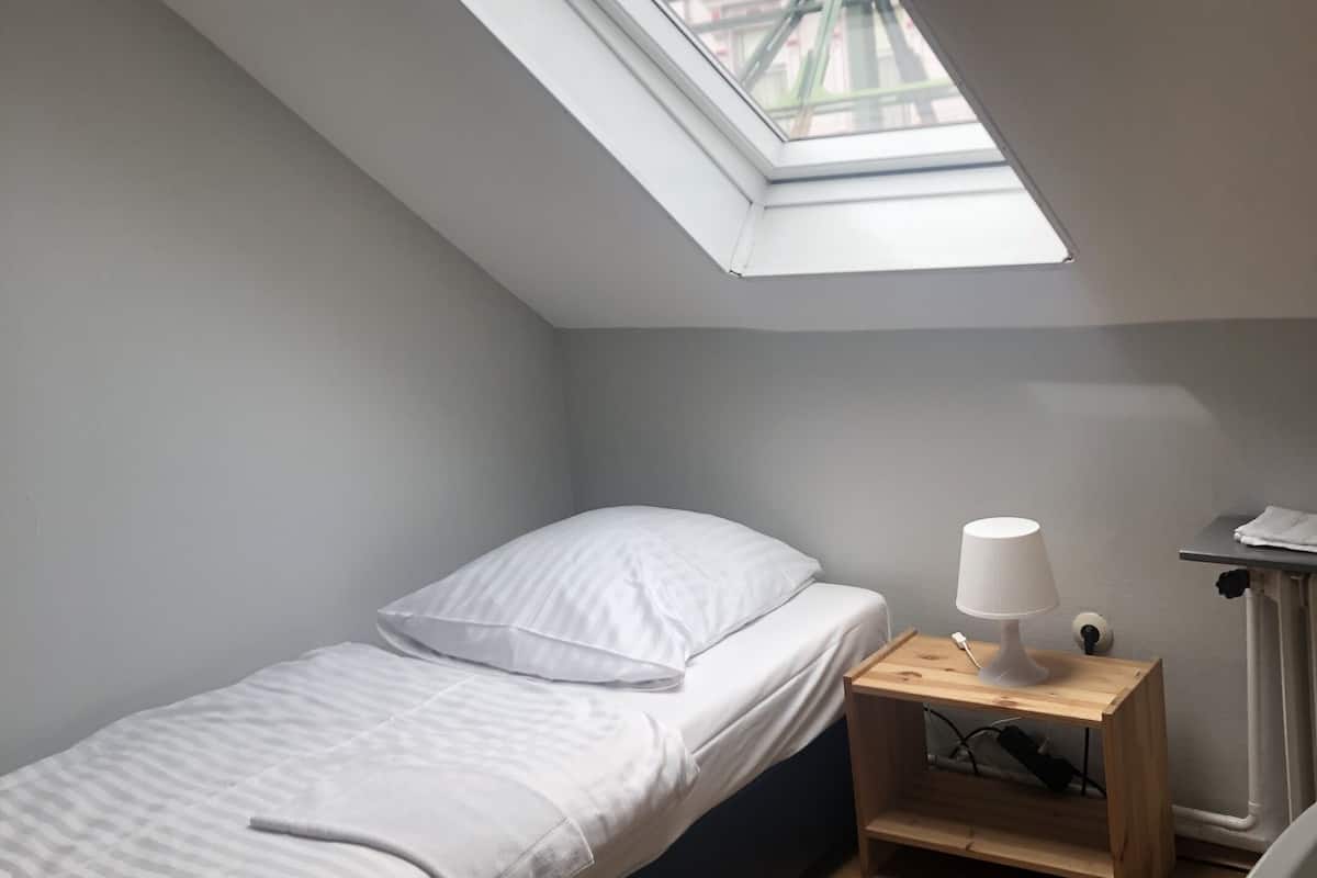 Quarto pequeno com uma cama de solteiro, janela no teto, mesa de cabeceira com abajur e uma cadeira.